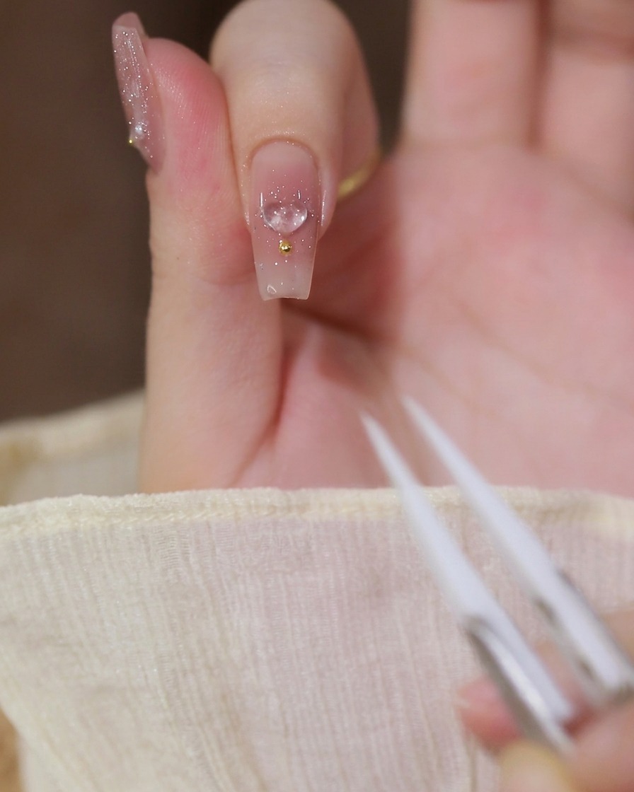 A close-up of an index finger with a milky nude gel base, featuring a clear 3D heart charm and a gold bead, with tweezers visible in the foreground.
