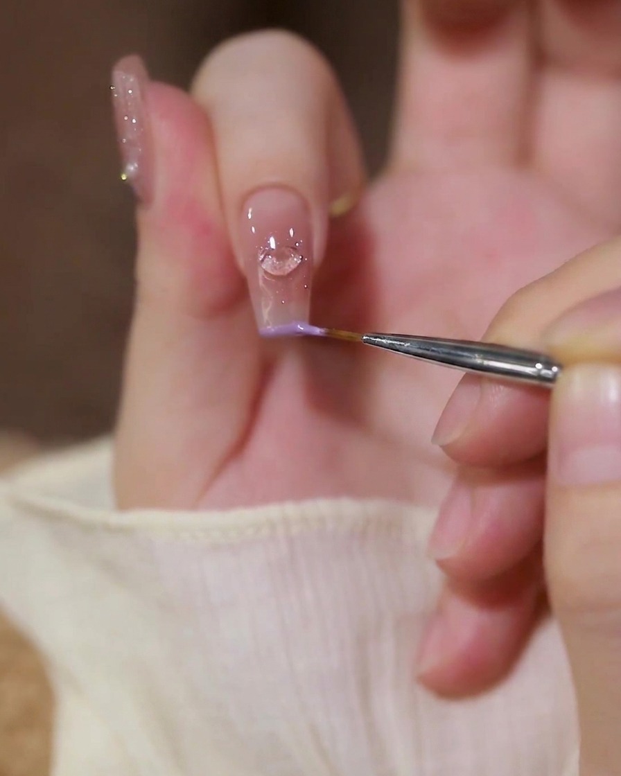 A close-up of a fine liner brush applying pastel purple gel polish along the edge of an index finger with a heart charm.