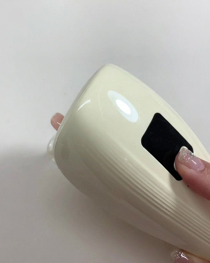 A hand holding a small white portable UV/LED lamp directly over a freshly painted nude gel nail to cure the polish.