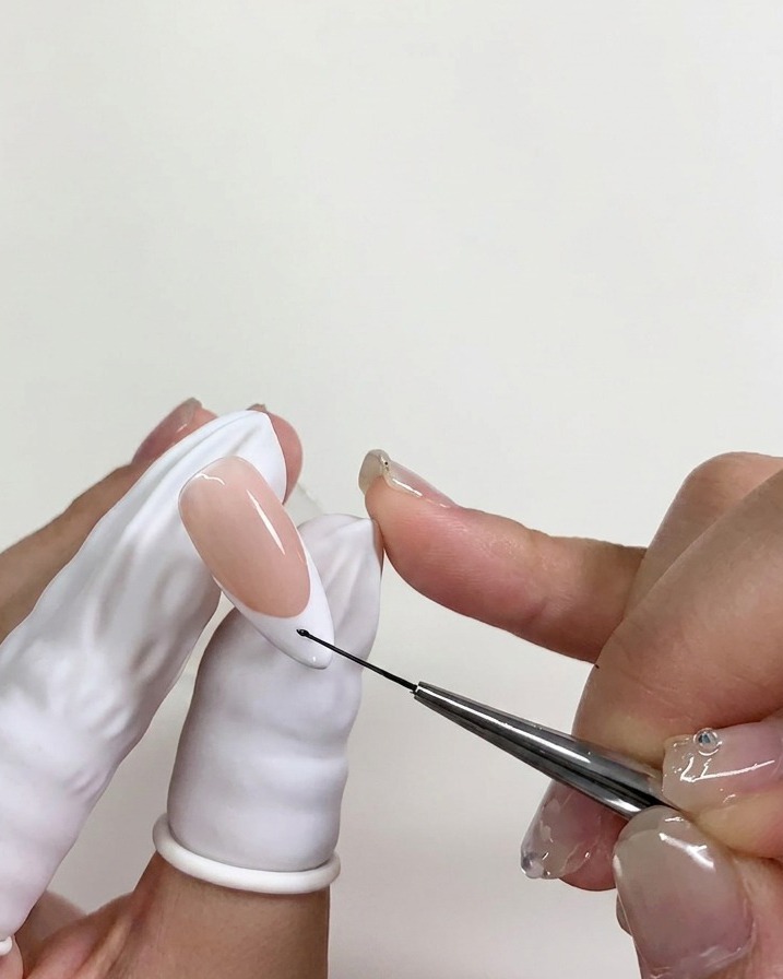 A fine nail art brush placing a tiny dot of black gel polish onto the white tip of a fingernail over a layer of uncured blooming gel.