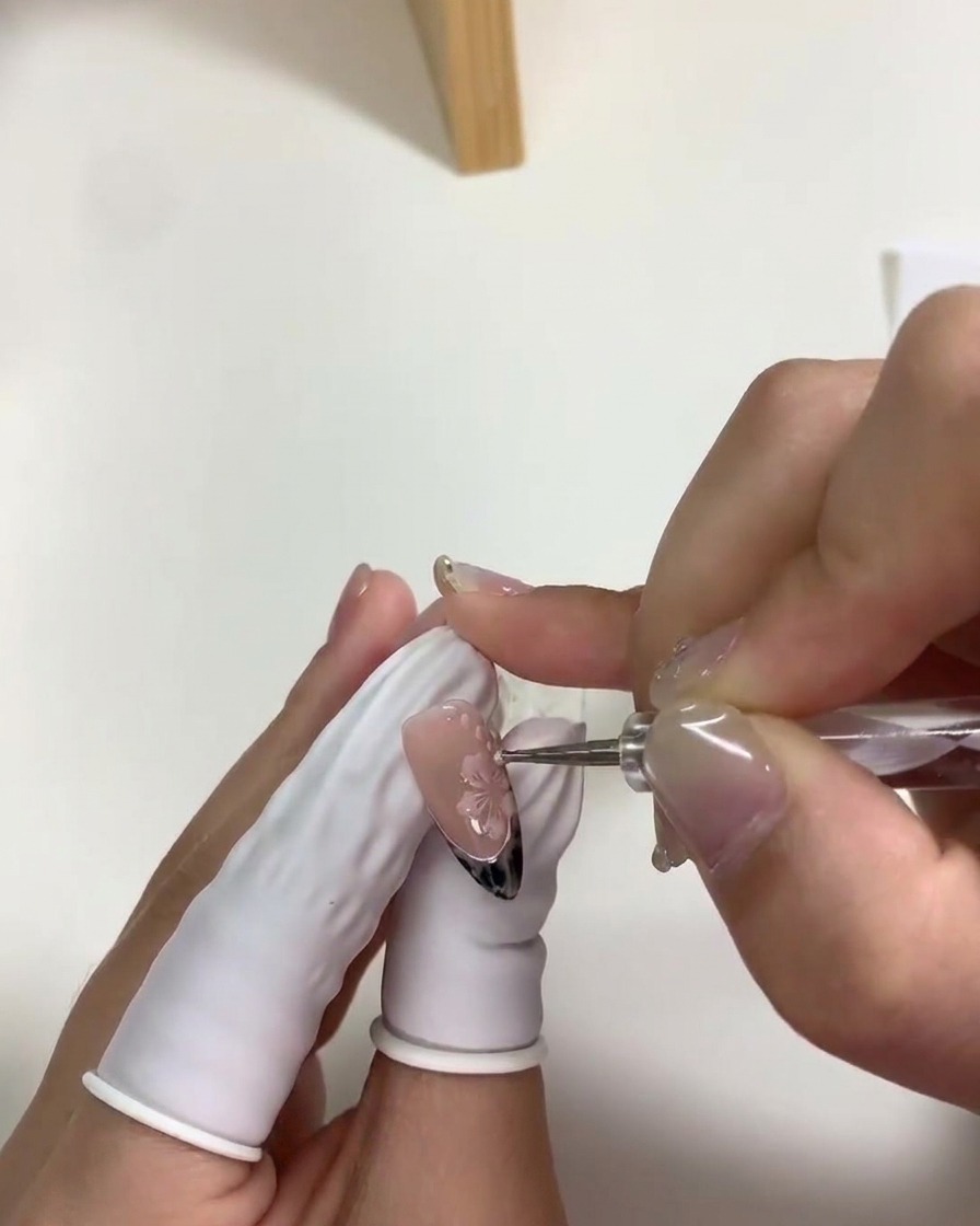A close-up view of a dotting tool placing small white accent dots around pink cherry blossoms on a fingernail.