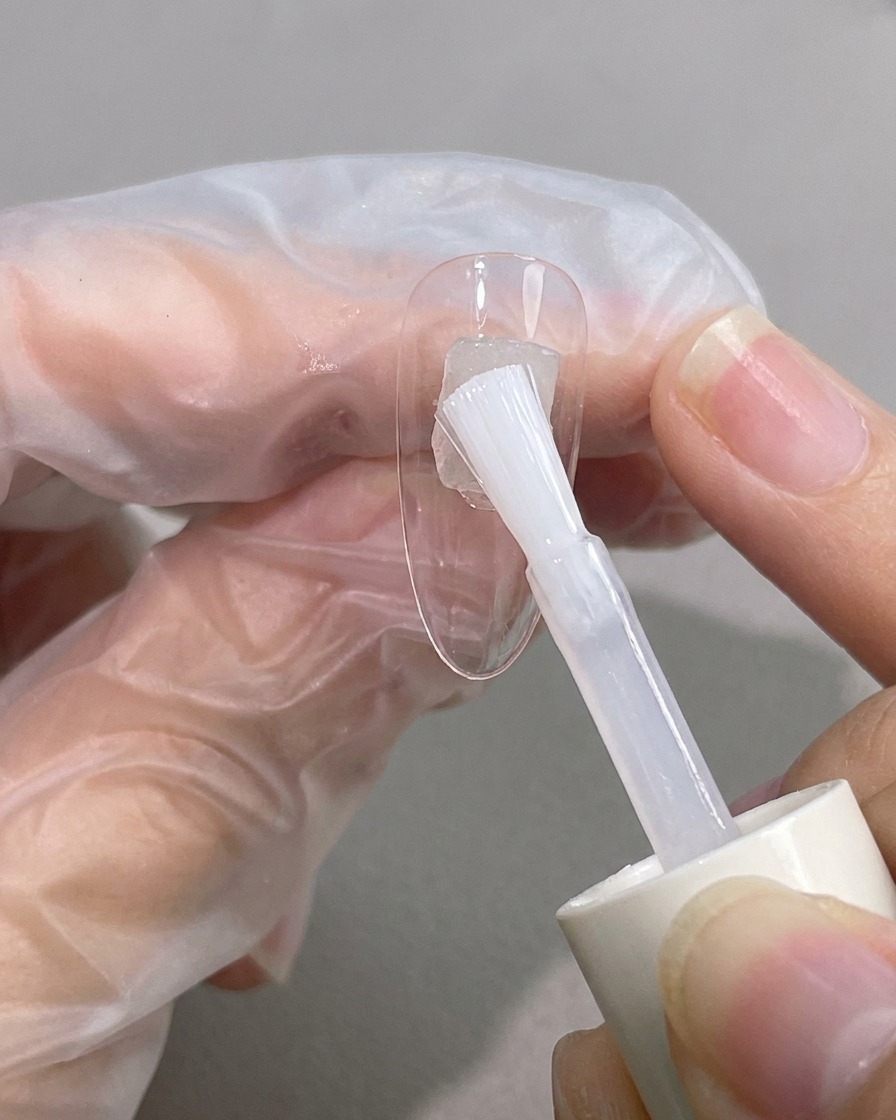 A brush applying a clear, viscous blooming gel diagonally across a natural nail tip.