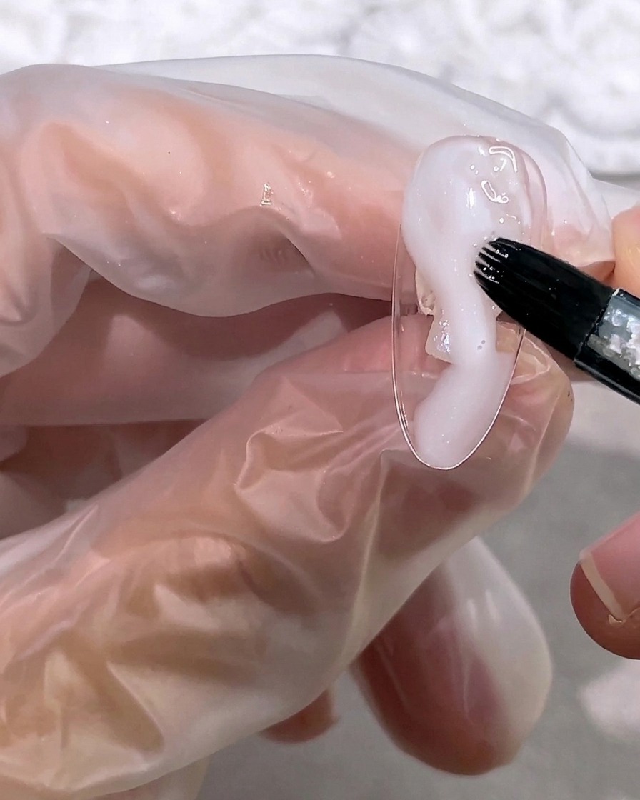 A brush applying a small amount of milky, semi-transparent white gel to the tip of a nail.