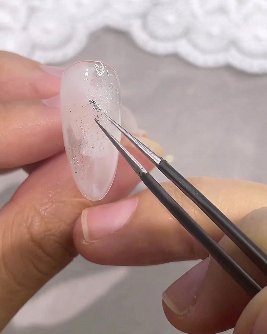 Tweezers placing a small, irregular piece of silver foil onto the nail.