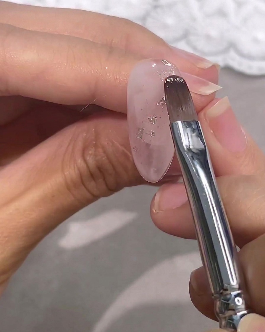 A brush applying a sheer pink gel polish over a clear nail with silver flakes.