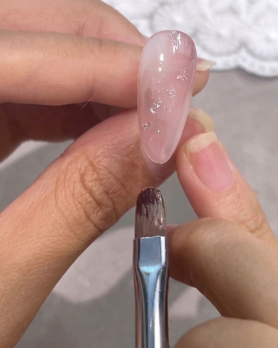 A detail brush hovering over a glossy pink gel nail with silver foil accents.