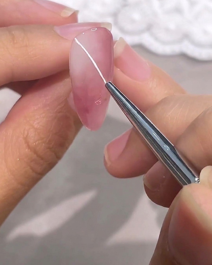 A fine detail brush drawing a thin white diagonal line across a pink gel nail.