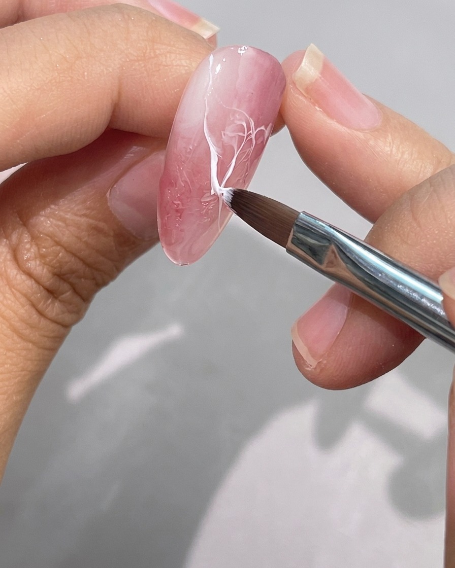 A fine detail brush blending and softening the edge of a white gel line on a pink marbled nail.