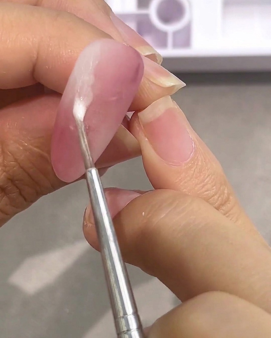 A small tool applying a drop of watery white paint onto a matte pink nail.