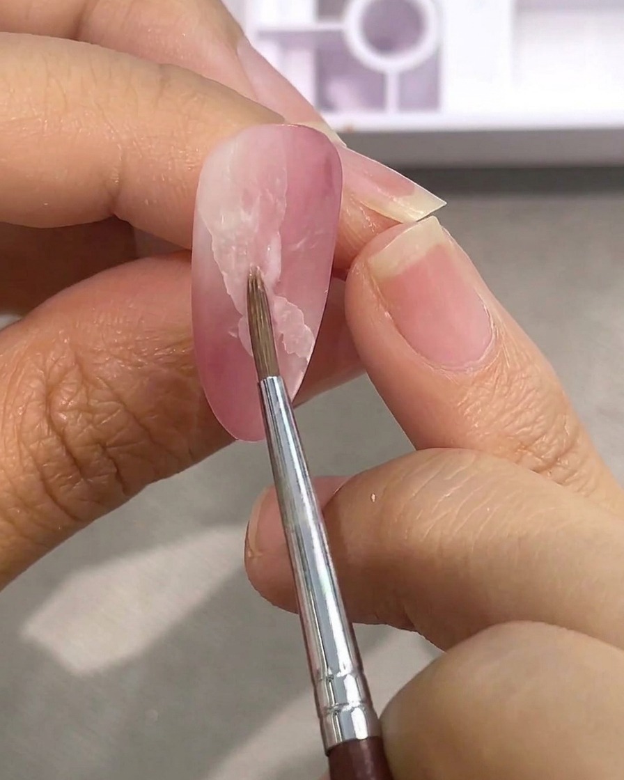A fine detail brush blending the edges of a white watercolor patch on a matte pink nail using water.