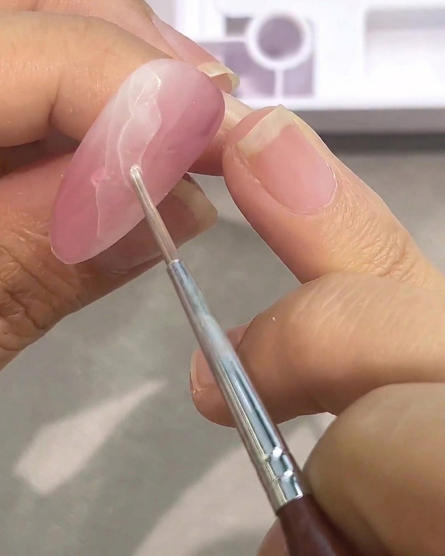 A close-up of a brush applying a second layer of white watercolor paint over a pinkish base, building up the marble pattern.