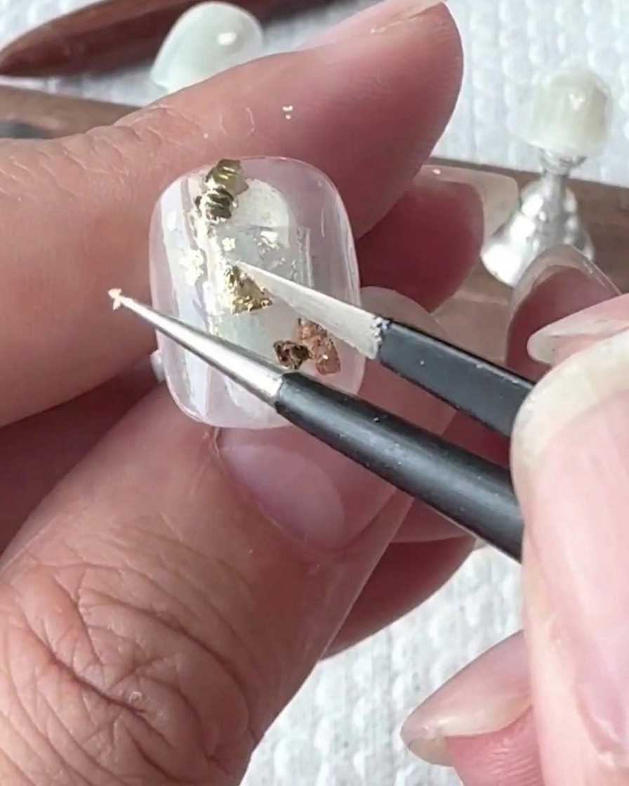 Tweezers carefully placing irregular pieces of gold foil onto the nail surface.