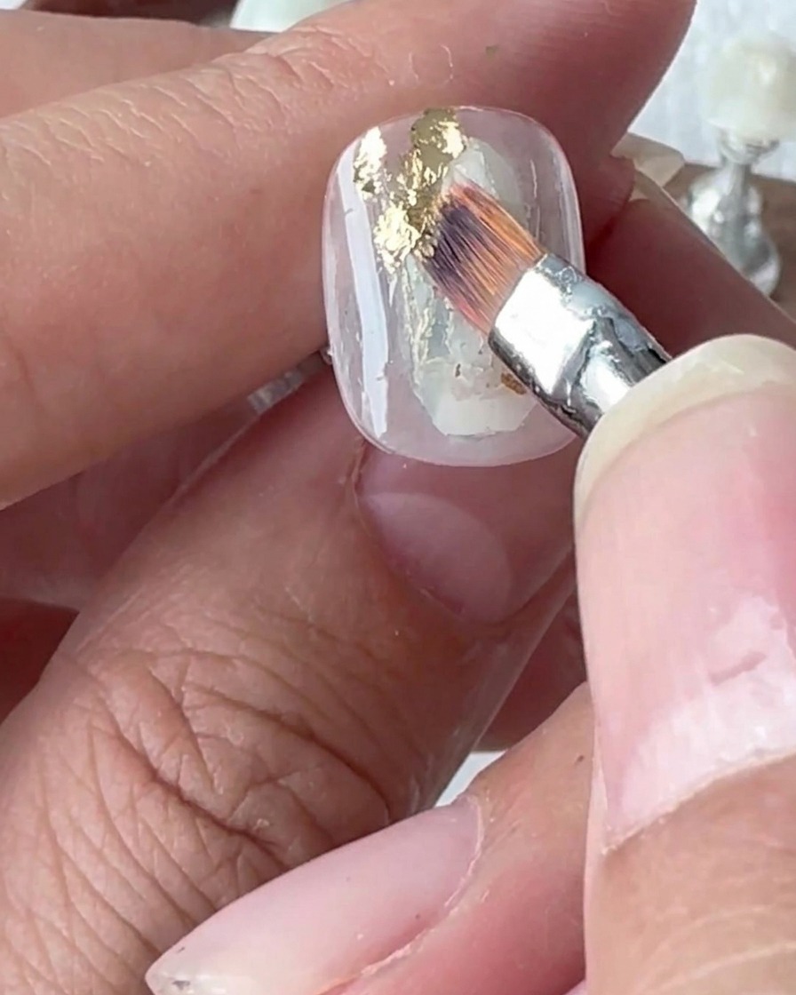 A close-up of a silicone tool pressing down onto gold foil flakes applied to a nail.