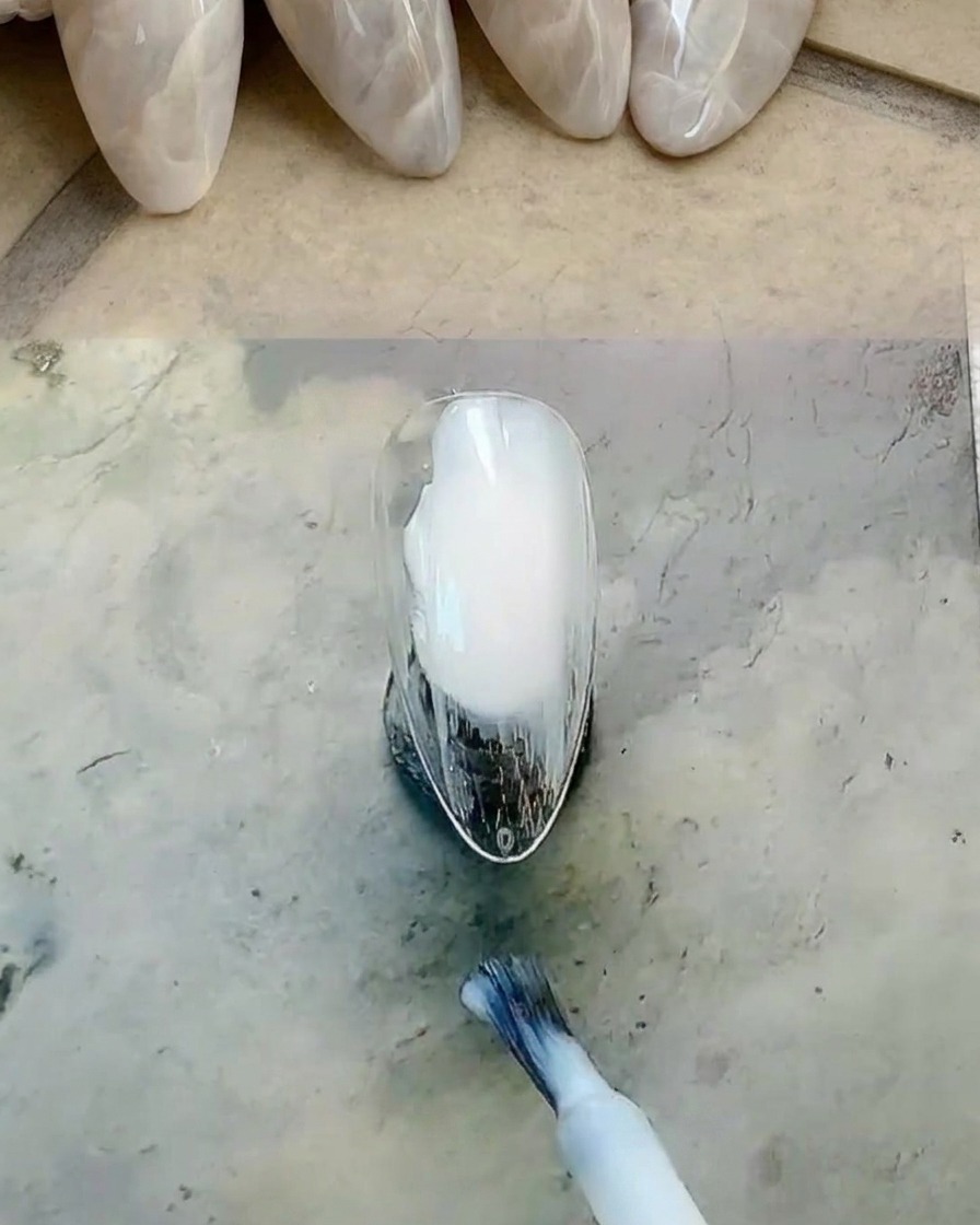 A bottle brush applying a milky white gel polish to a nail surface.