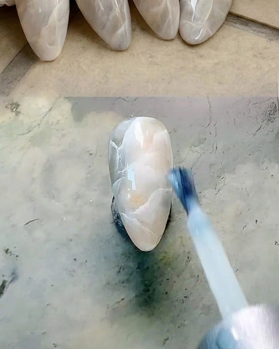 A flat brush applying a thin coat of translucent milky white gel polish over a marbled nail.