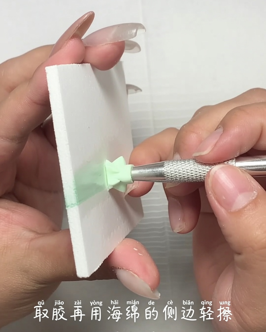 A close-up view of a metal tool applying a small amount of light green gel polish from a palette onto the edge of a makeup sponge.