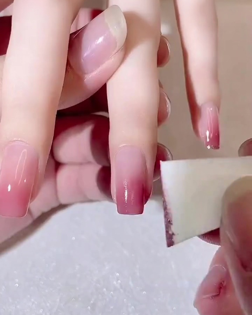 A close-up view of a makeup sponge being used to dab a second layer of dark pink gel onto a nail tip.