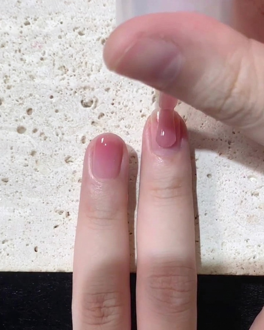 A close up of a nail brush applying a sheer pink jelly polish near the cuticle area of a natural nail