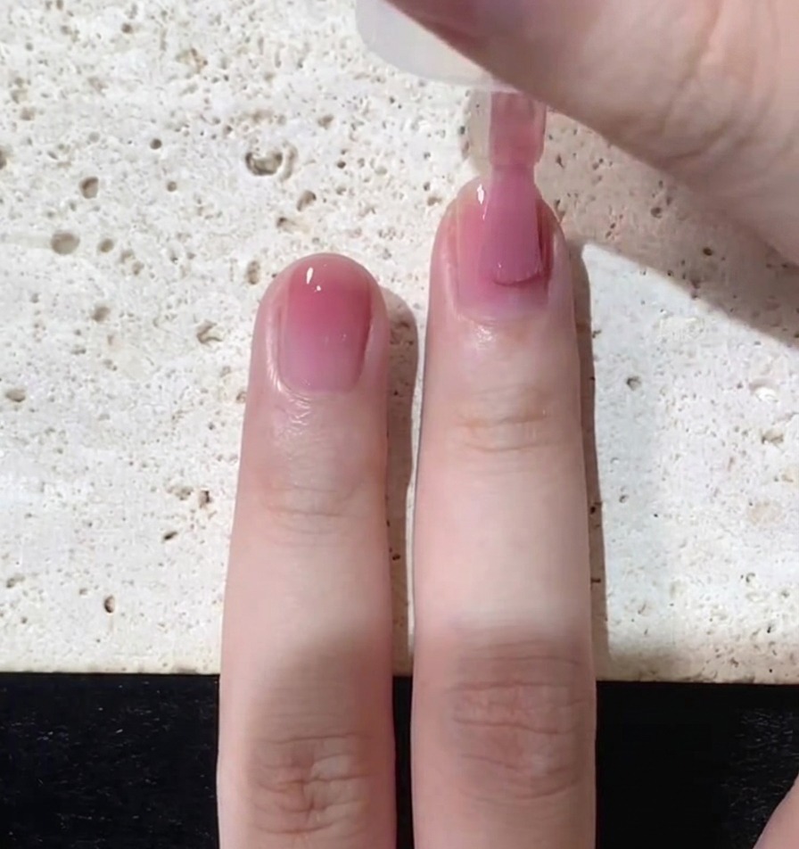 A close up shot of a nail brush applying a thin layer of translucent pink polish over a natural nail