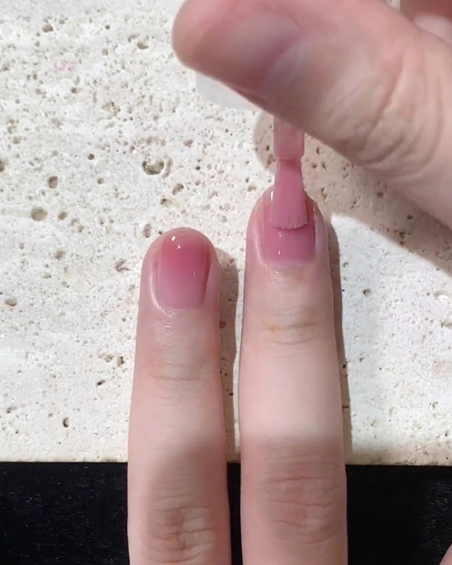 A nail brush sweeping pink jelly polish smoothly down towards the free edge over the top half of the nail.