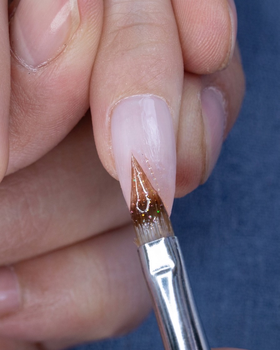 A close-up view of a fine brush applying small, colorful glitter particles onto the tip of a nude-pink ombre nail.