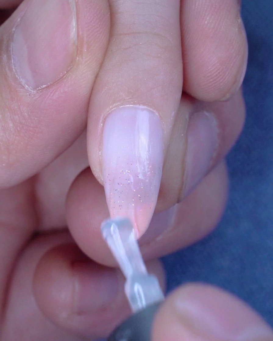 A brush applying a smooth, clear layer of glossy top coat over a glitter-accented ombre nail.
