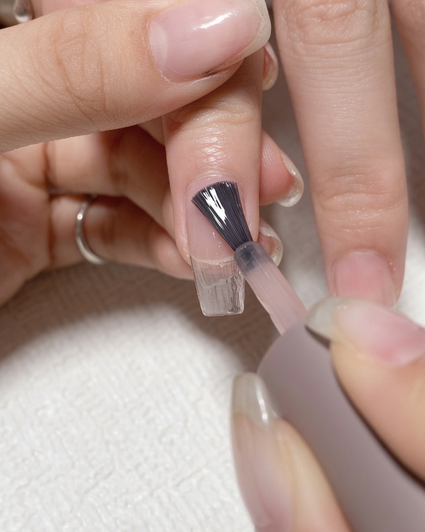 A brush applying clear base coat gel to a clean natural fingernail.