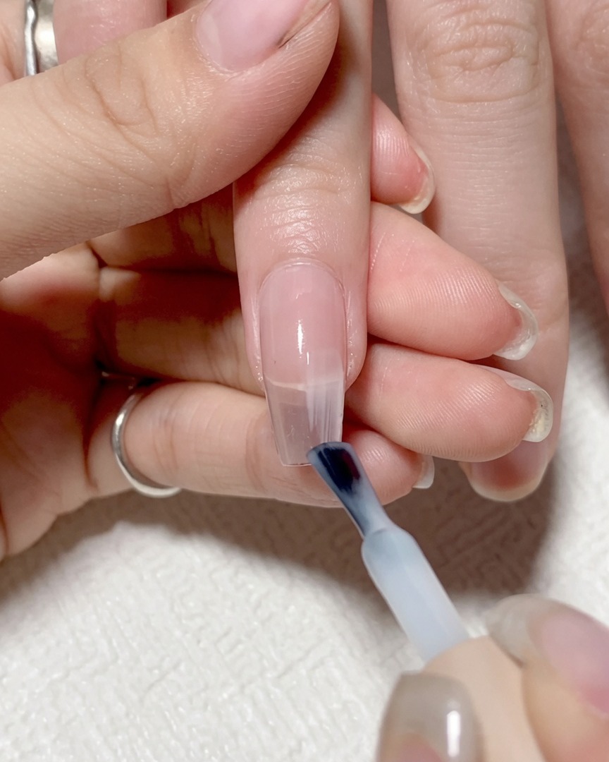 A nail brush applying sheer, light-colored gel to the right side of a nail.