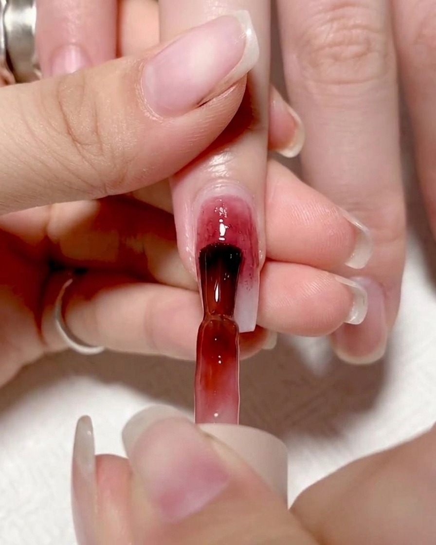 A brush applying a vibrant red gel to the left half of a fingernail, creating a distinct color block.