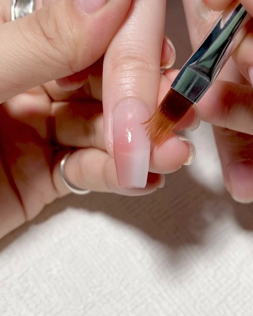 A brush applying a clear, slightly thick strengthening gel over the full nail surface.