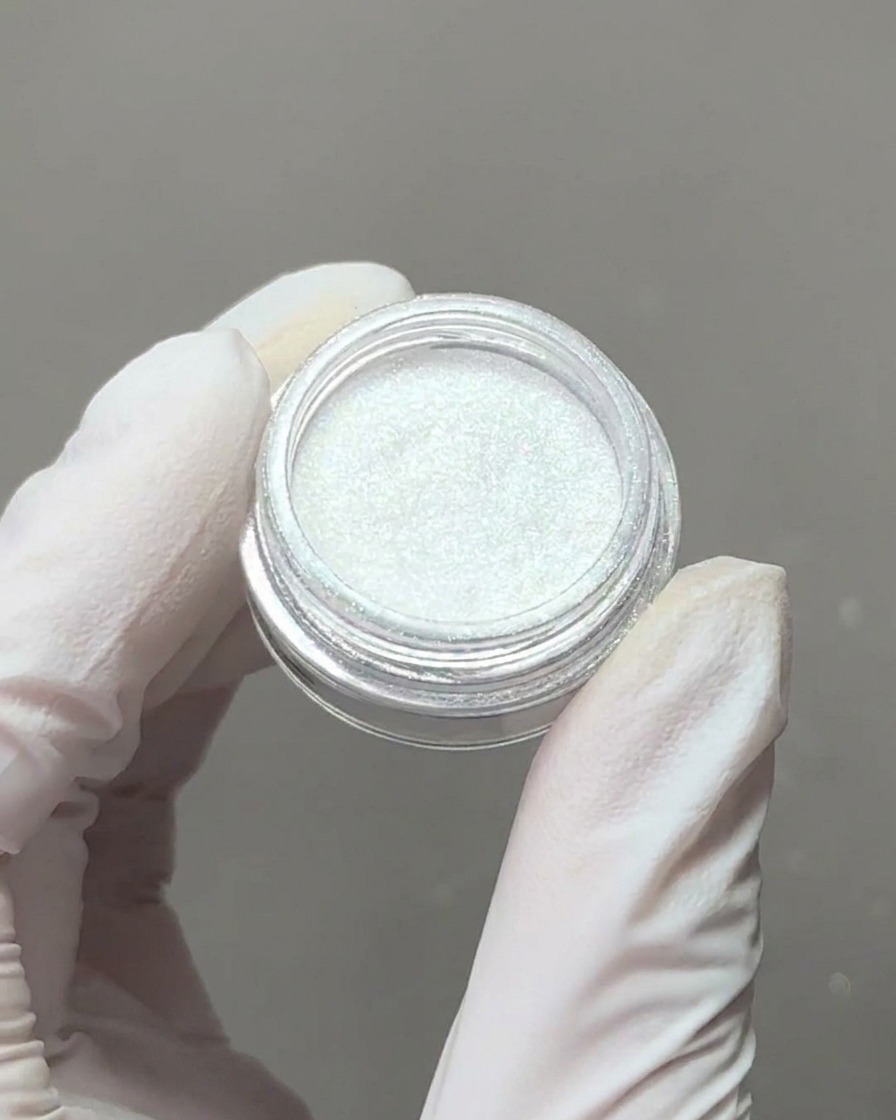 A gloved hand holding a small container of iridescent aurora chrome powder with a sponge applicator tip picking up some of the pigment.