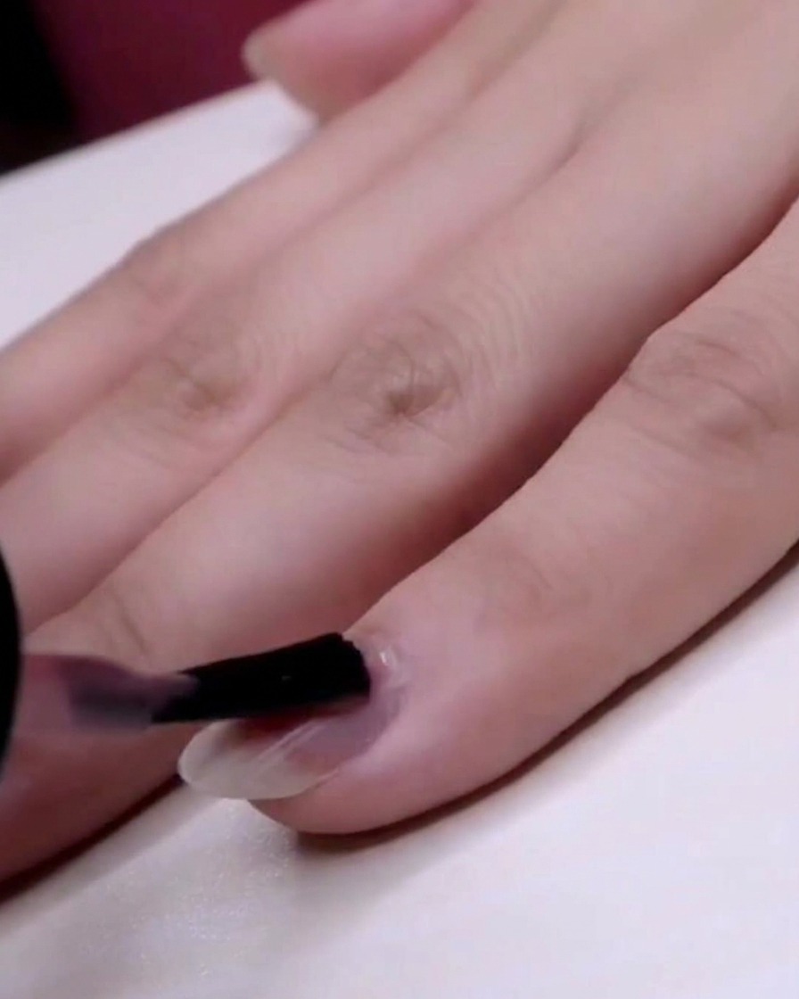 A brush applying a transparent pink jelly polish over the full extent of a pinky nail.
