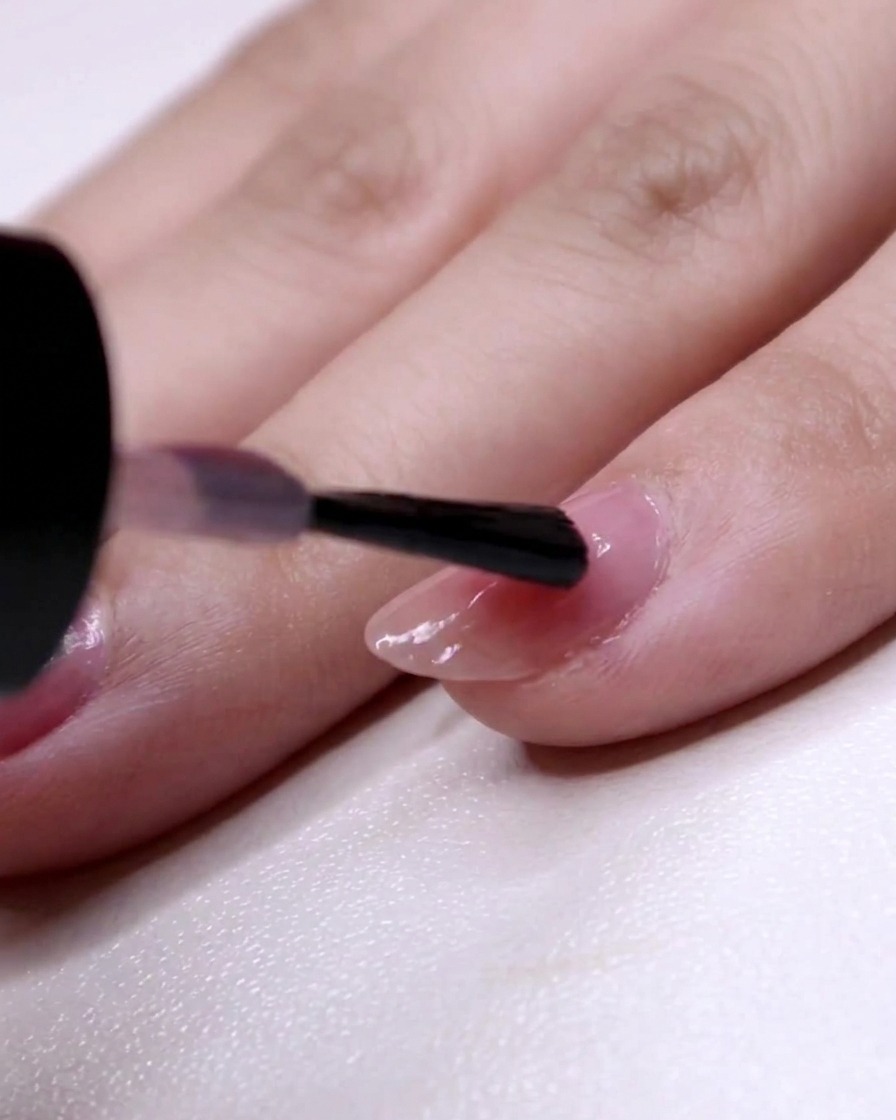 A nail brush painting the second layer of pink polish from the two-thirds position to the ring finger tip.