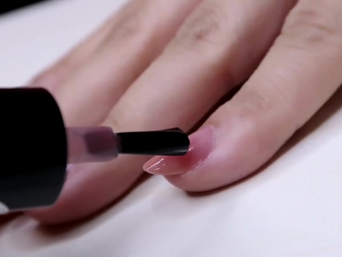 A brush applying a coat of sheer pink polish to the outer half of a fingernail.
