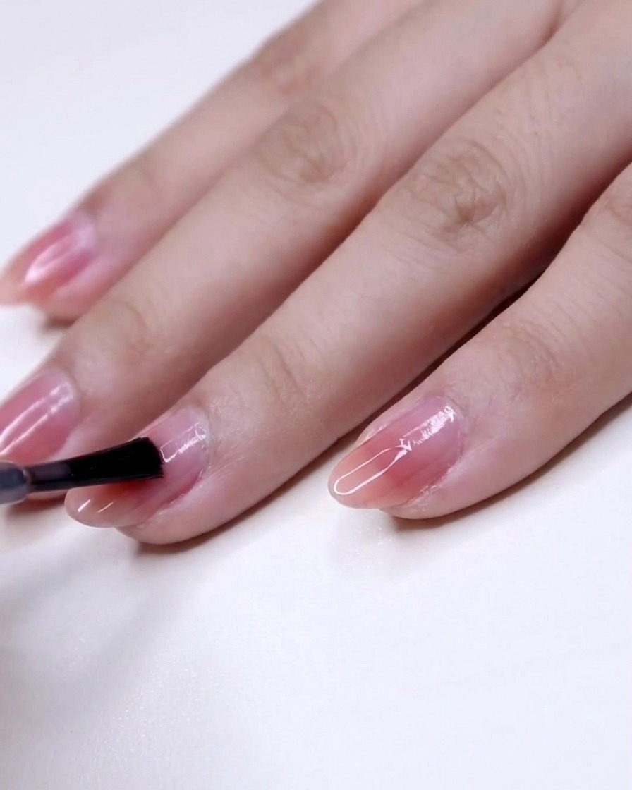 A close-up of a brush applying pink polish from the center to the tip of a fingernail.