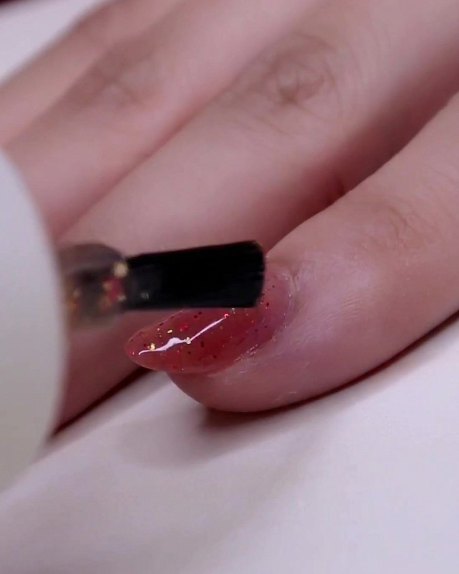 A close-up of a brush applying glitter polish to the ring finger, focusing on placing gold flakes securely.