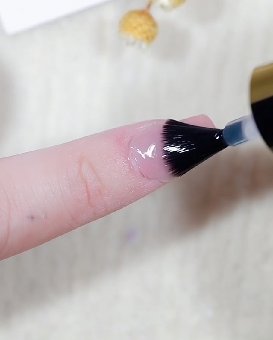 A close-up of a brush applying clear top coat over a nail that has a shimmering aurora effect.