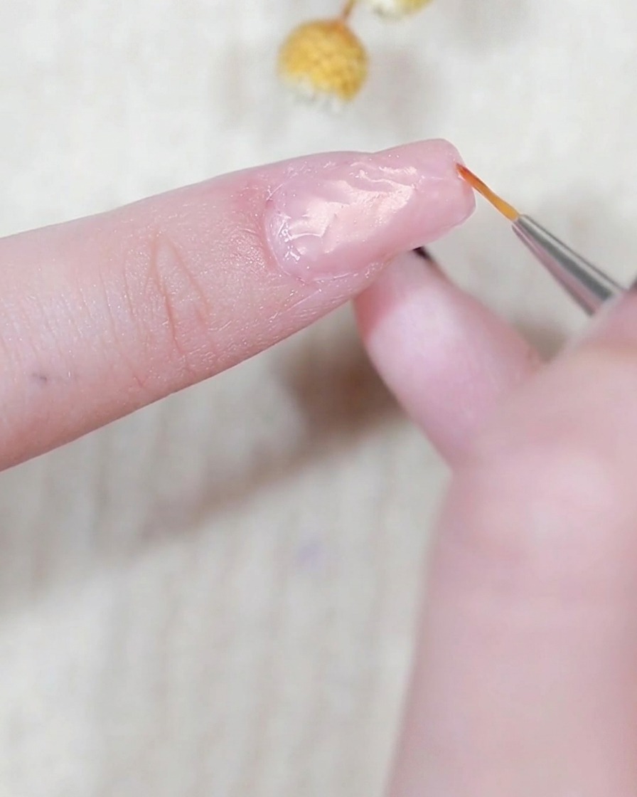 A detail shot of a fine liner brush applying a thin, raised line of clear modeling gel around the perimeter of a nail.