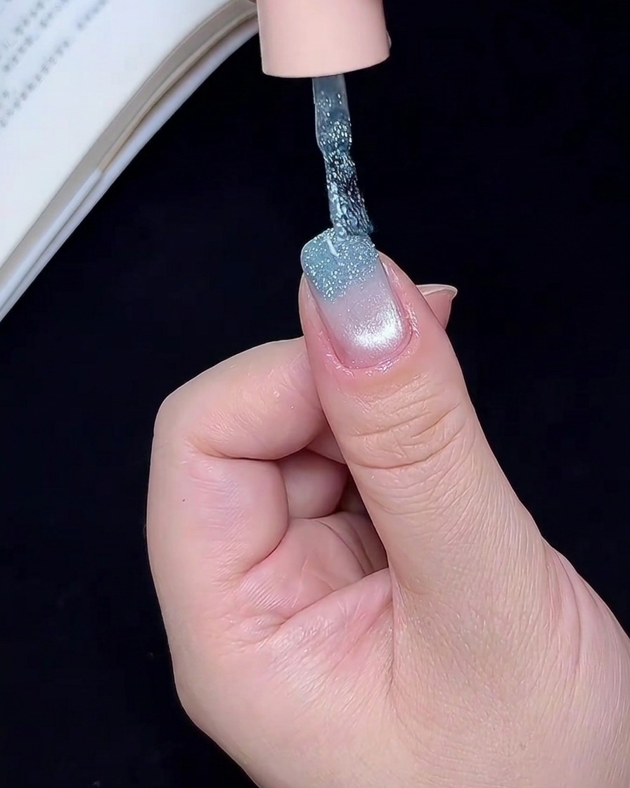 A nail art brush applying shimmering blue glitter gel to the free edge of a fingernail.