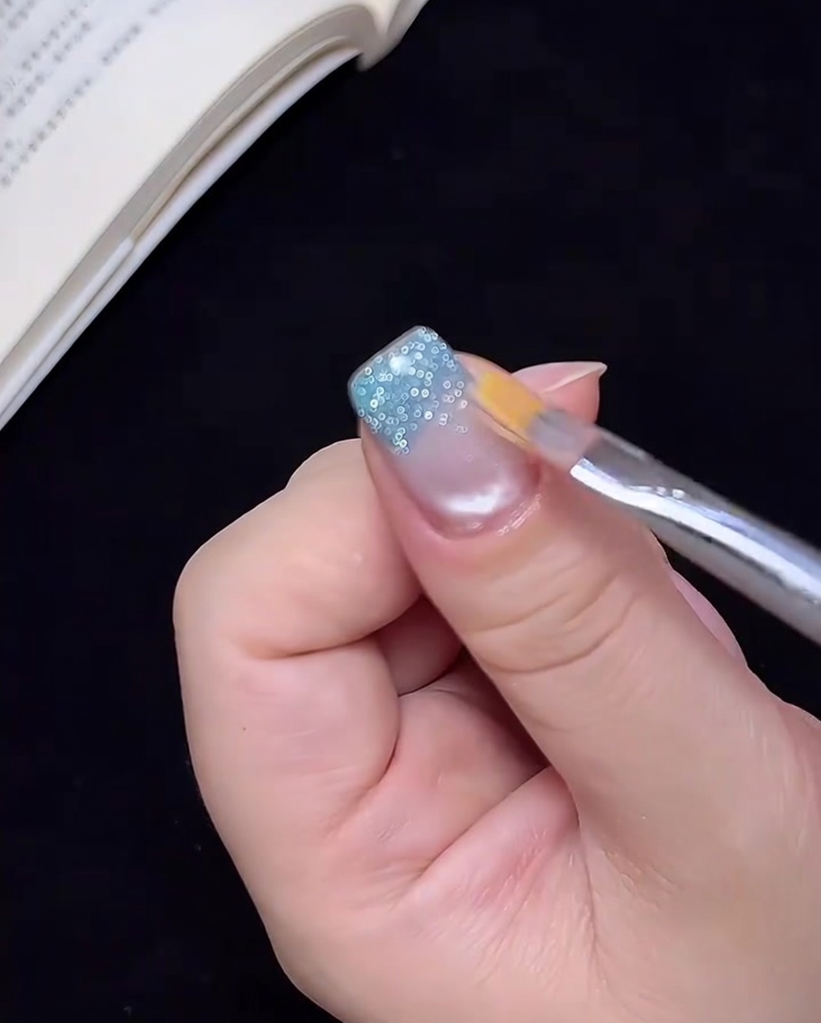 A clean brush lightly dragging blue glitter gel upward on a fingernail to create an ombre effect.
