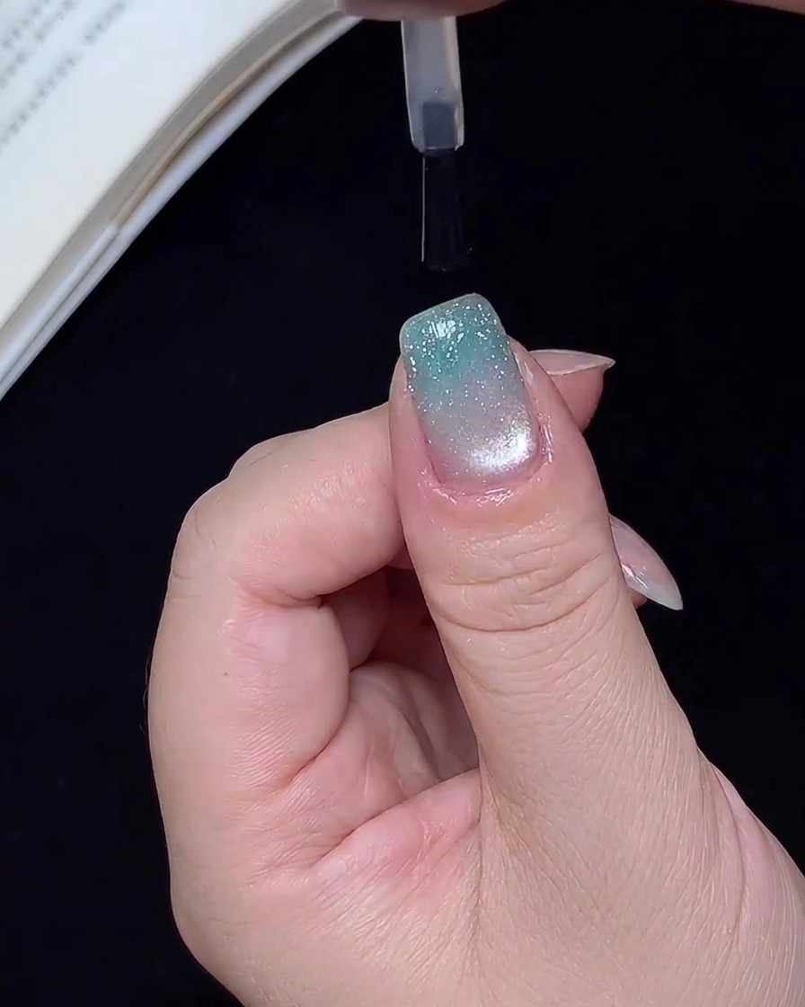 A brush applying a clear, glossy top coat over a finished blue glitter gradient nail.