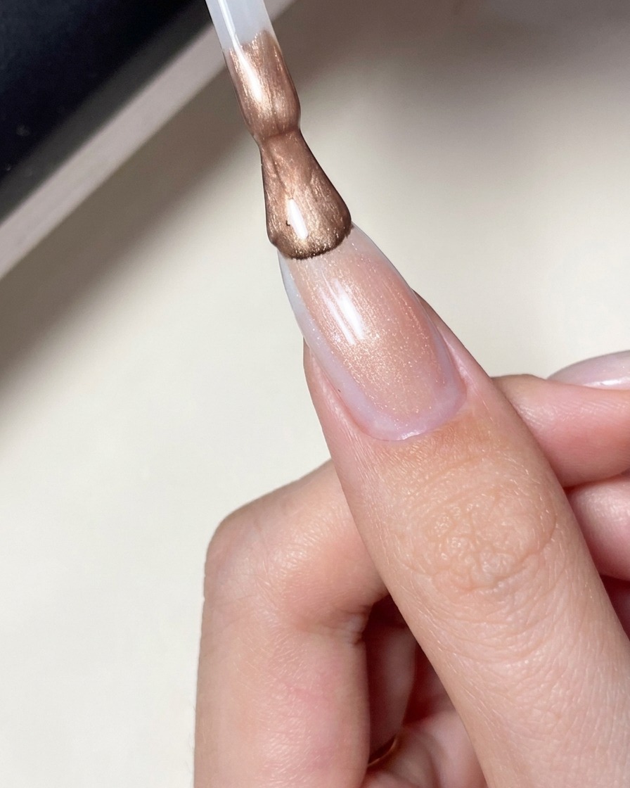 A brush applying a sheer, milky pink gel polish over a nail with a star design.