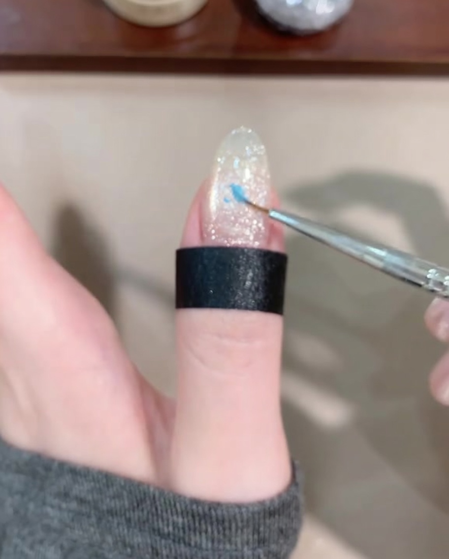A fine liner brush applying a small drop of blue gel to the center of the nail.