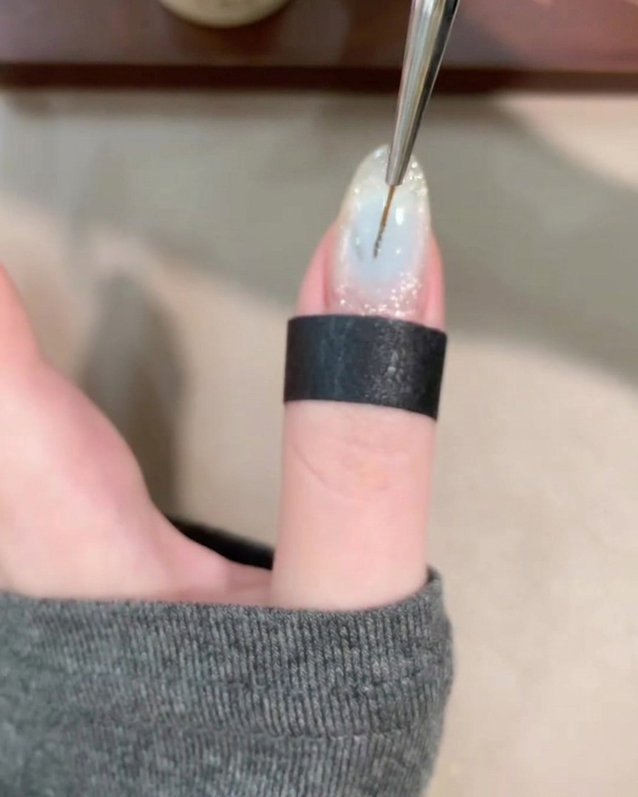 A close-up view of a thin liner brush drawing fine, swirling lines over a clear gel base on a fingernail.