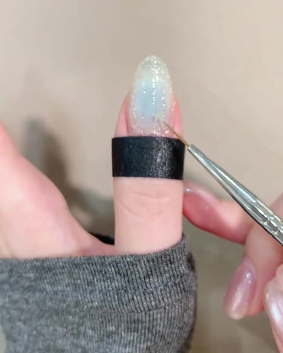 A pair of tweezers placing a small, shiny gold star-shaped sequin onto a clear nail decorated with glitter and faint blue tones.
