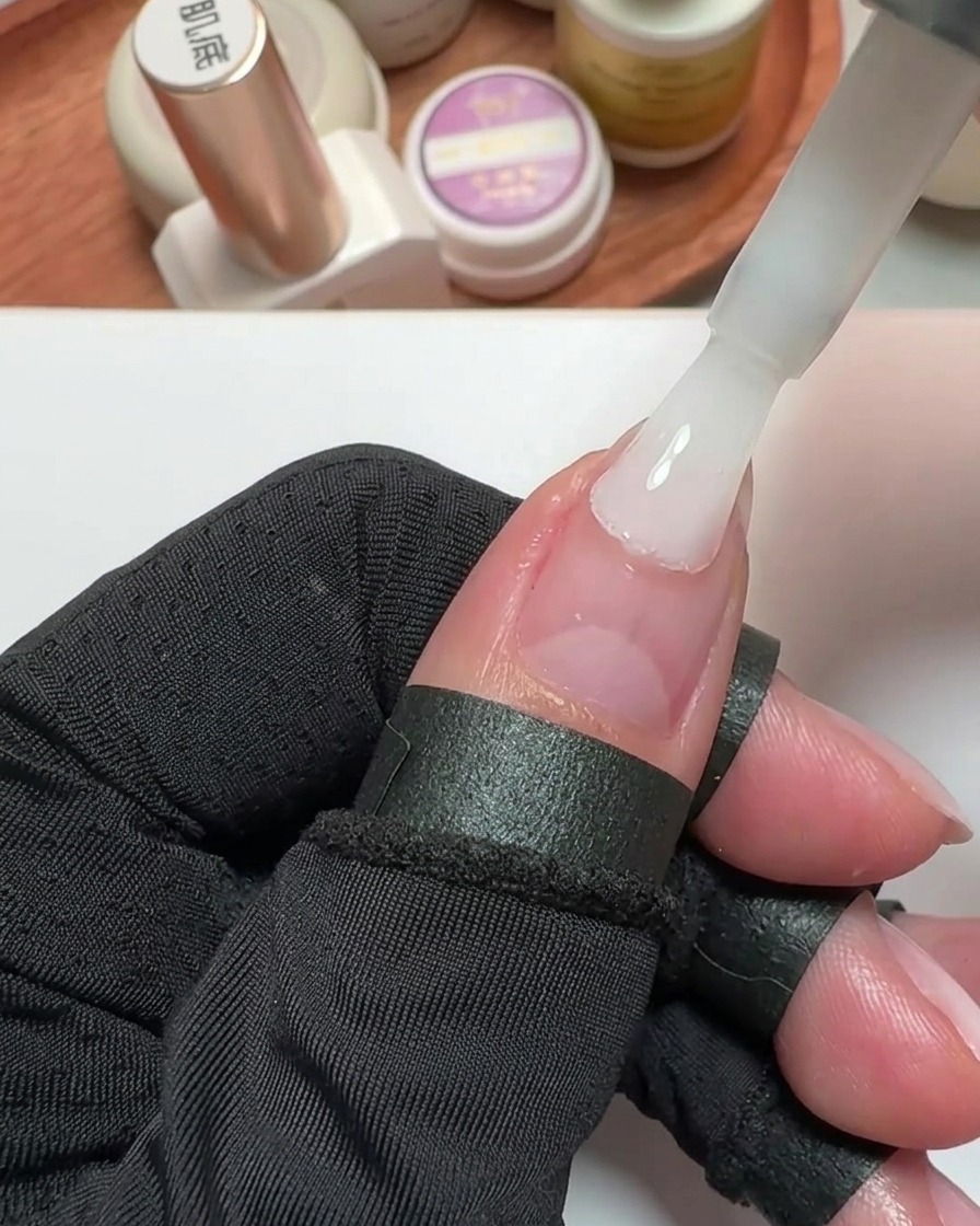 A close-up of a brush applying milky white gel polish to a prepared thumb nail.