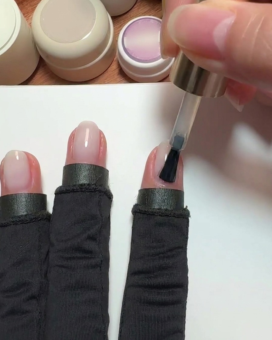 A brush applying a sheer pink nude gel polish to an index fingernail.
