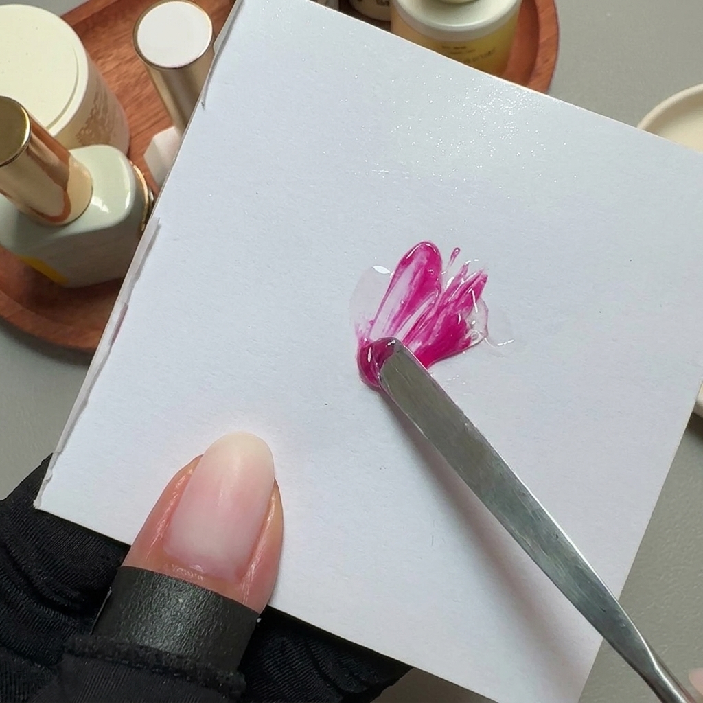 A metal spatula mixing bright pink gel polish on a white square mixing palette.