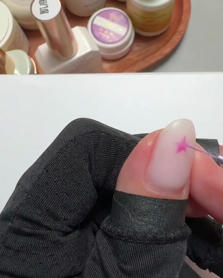 A close-up of a fine liner brush painting a bright pink star shape on a fingernail with a milky white base.