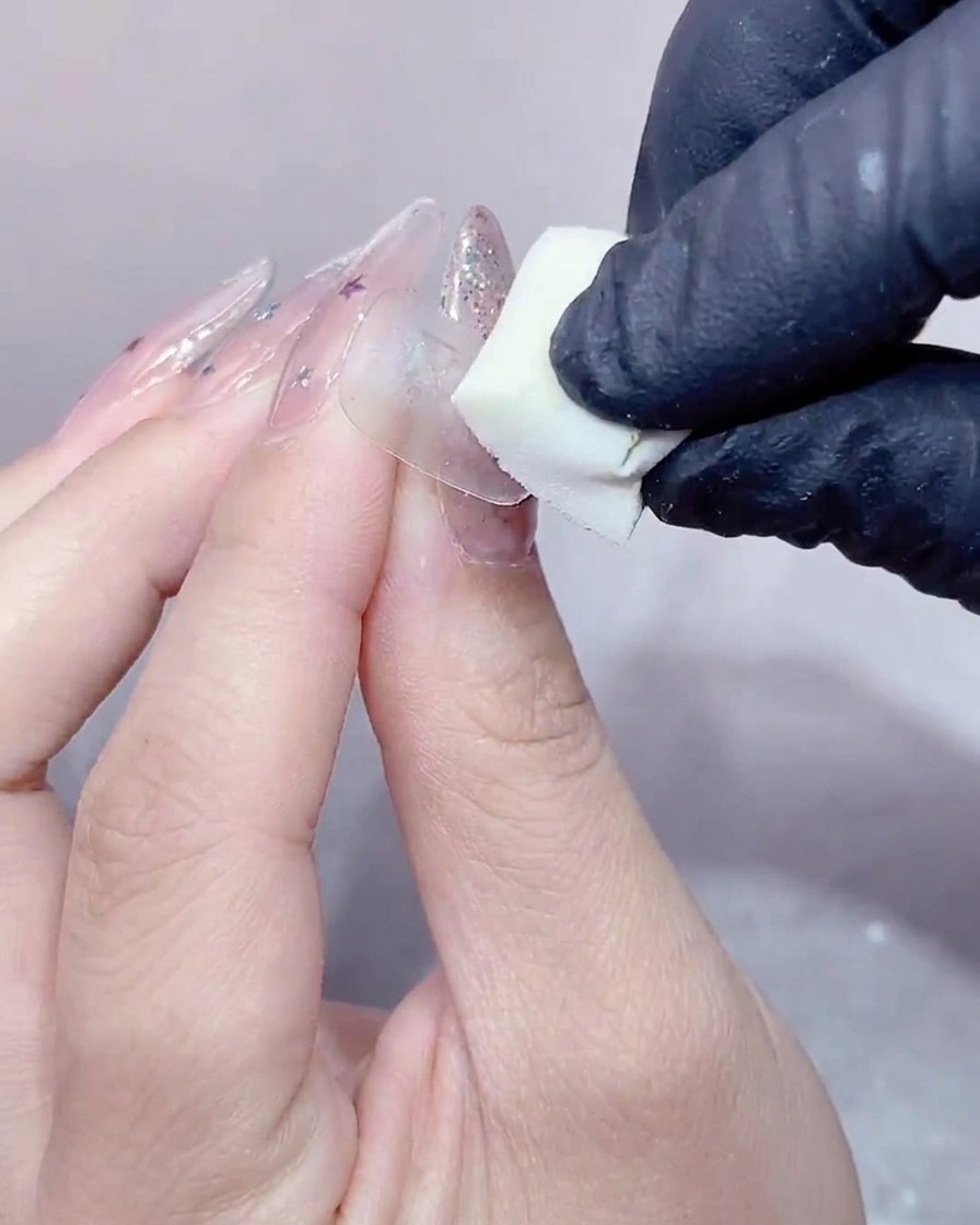 A gloved hand pressing a small white makeup sponge onto the surface of a clear gel-covered nail.